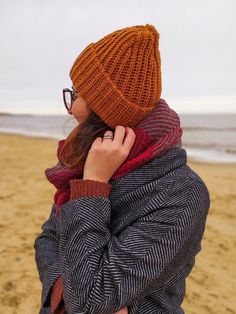 a woman wearing glasses and a knitted hat is standing on the beach while talking on her cell phone