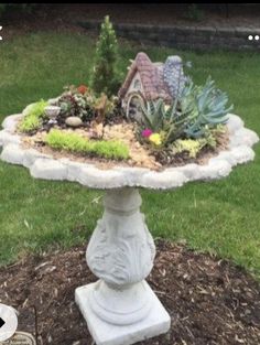 a bird bath with plants and rocks in the ground next to a house on a lawn