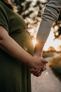 a pregnant woman holding the hand of her husband as they walk down a path at sunset