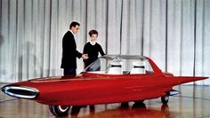 two men shaking hands in front of a red car that is on display at an exhibition