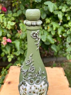 a green vase sitting on top of a wooden table in front of some bushes and flowers