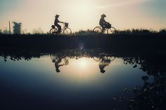 two people riding bikes on the side of a river at sunset or dawn with their reflection in the water