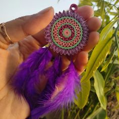 a hand holding a purple and green brooch with feathers in front of some plants