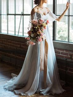 a woman standing in front of a window wearing a wedding dress and holding a bouquet