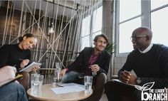 three people sitting at a table with drinks and papers in front of them, talking to each other