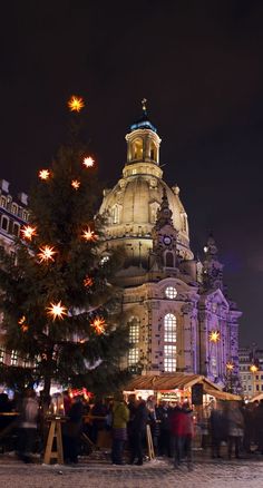 people are gathered around the christmas tree in front of a large building with lights on it