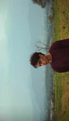 a young man standing in front of a body of water wearing a maroon shirt and black pants