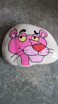 a painted rock with a pink tiger on it's face and yellow eyes is sitting on the ground