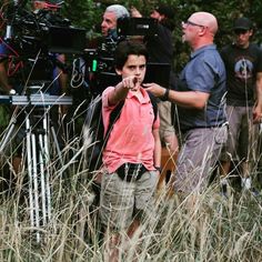 a young boy standing in tall grass next to a camera set up on a tripod