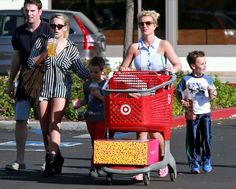 a woman pushing a red shopping cart with two children and an adult walking behind her