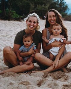 three women and two babys sitting in the sand at the beach with trees in the background