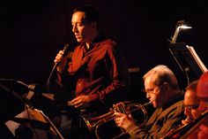 two men playing musical instruments in front of a microphone and another man holding a microphone