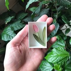 a hand holding a small square glass object with pink flowers on it and green leaves in the background