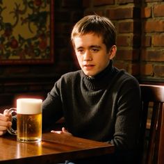 a young man sitting at a table with a mug of beer in front of him