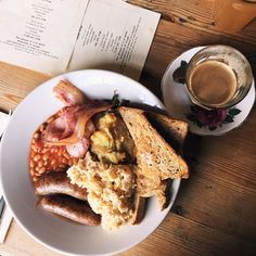 a white plate topped with lots of food next to a cup of coffee and an open book