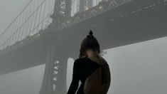 a woman standing in front of the brooklyn bridge on a foggy day with her back to the camera