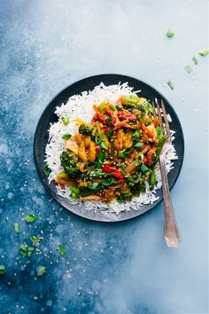 a black plate topped with rice and veggies next to a fork on a blue surface