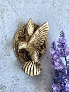 a gold bird brooch sitting on top of a wall next to purple and white flowers