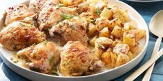 a white plate topped with chicken and potatoes next to two silver spoons on a blue table cloth