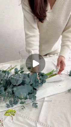 a woman is arranging flowers on a table with white linens and greenery in front of her