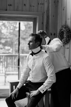 two men in formal wear are sitting on a bench and one man is adjusting his bow tie