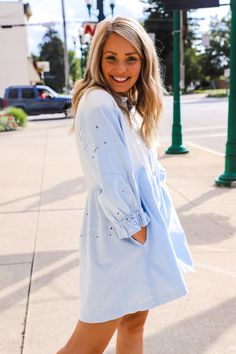 a woman is standing on the sidewalk in her blue shirt dress and tan shoes, smiling at the camera