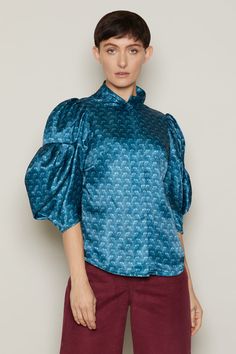 The Salvar is a shimmering shirt in two colourways of Mountain Brown and Shore Blue, influenced by Icelandic fjords and valleys.An iridescent satin feel with extravagant balloon sleeves to the elbow, the Salvar features a high ruffle neck and press stud fastenings.Haute couture and sustainability align in the Salvar shirt through the use of 100% polyester, vegan, roll end, designer fabric. Inclusive sizing, available in XS to 6XL.Each blouse is numbered and production is limited to 100 pieces pe Blue Long Sleeve Satin Top, Blue Silk Blouse With Blouson Sleeves, Elegant Blue Blouse With Gathered Sleeves, Blue Satin Long Sleeve Top, Blue Fitted Blouse With Balloon Sleeves, Silk Tops With Blouson Lantern Sleeves, Silk Tops With Lantern Sleeves, Party Tops With Gathered Balloon Sleeves, Evening Tops With Gathered Balloon Sleeves