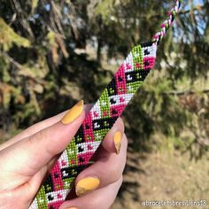 a hand holding a colorful beaded object in front of some trees and bushes with yellow nails