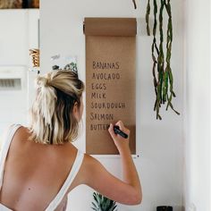 a woman writing on a sign in front of a wall with plants hanging from it