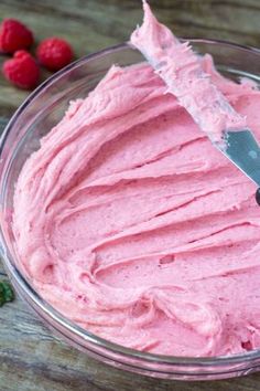 a bowl filled with pink frosting next to raspberries on a wooden table