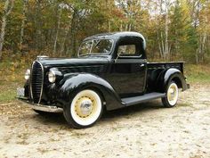 an old black truck is parked on the dirt in front of some trees and leaves