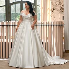 a woman in a white wedding dress standing next to a wooden wall and window with her hands on her hips