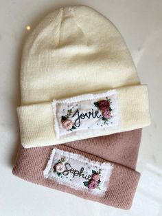 two beanies with embroidered name tags on the front and back, one is light pink