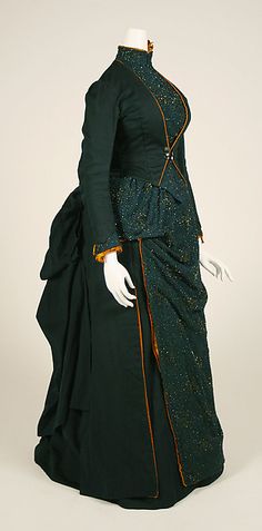 American gown (circa 1887).  Silk & wool dress, with trimmings of glass beading & feathers. The solid colored dark teal wool forms the cut away jacket and is tucked into a bustle at the back of the gown.