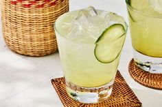 two glasses filled with lemonade and cucumber on top of coasters next to wicker baskets