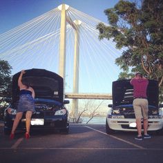 two people sitting on the hoods of their cars in front of a suspension bridge