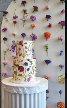 there is a white cake with flowers on the wall and behind it are purple, red, yellow and orange flowers