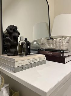 a stack of books sitting on top of a white dresser next to a lamp and mirror