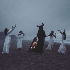 five women dressed in white dancing around a campfire on the beach at night time