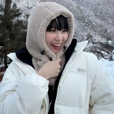 a woman wearing a white jacket and black hoodie smiles at the camera while standing in front of snow covered trees