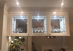 a kitchen with white cabinets and glass windows above the counter top, in front of a vase filled with flowers