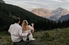 a man and woman sitting on top of a hill looking at the mountains with drinks in their hands