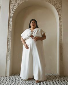 a woman standing in front of an archway with her hands on her hips, wearing a white jumpsuit