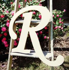 a white wooden sign with the letter r on it in front of flowers and bushes