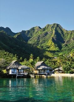 some huts that are sitting on the water near trees and mountains in front of them