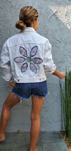 a woman standing next to a wall wearing a jean jacket with flowers painted on it