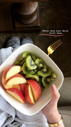 a person holding a white bowl filled with sliced kiwis and apples next to a fork