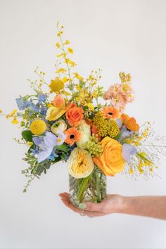 a hand holding a vase filled with lots of different colored flowers on top of a white wall