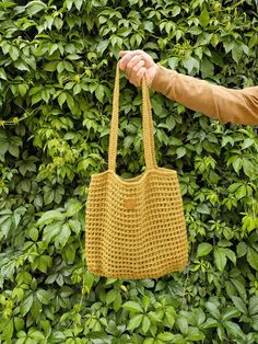 a hand holding a yellow crocheted purse in front of green bushes and shrubbery