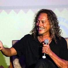 a man with long hair sitting in a chair holding a microphone and giving a speech